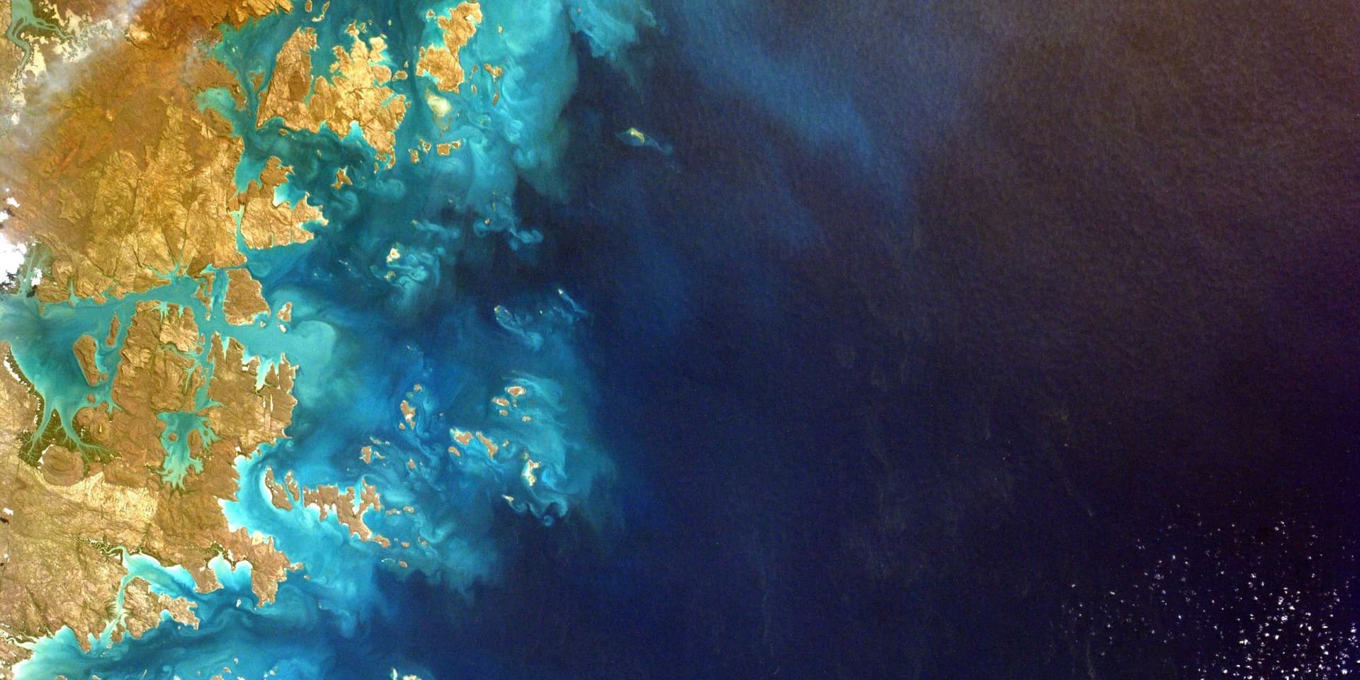 Vista de satélite de um litoral, com bancos de areia claros dissolvendo-se na água azul-profunda