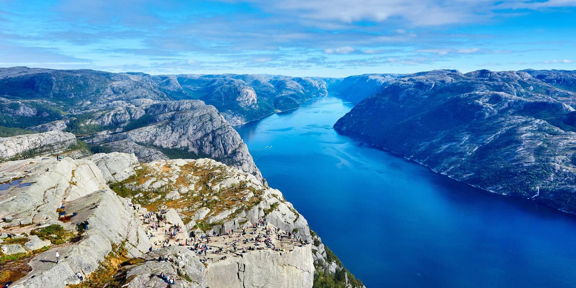Vista do alto de um penhasco sobre um fiorde azul cercado por montanhas