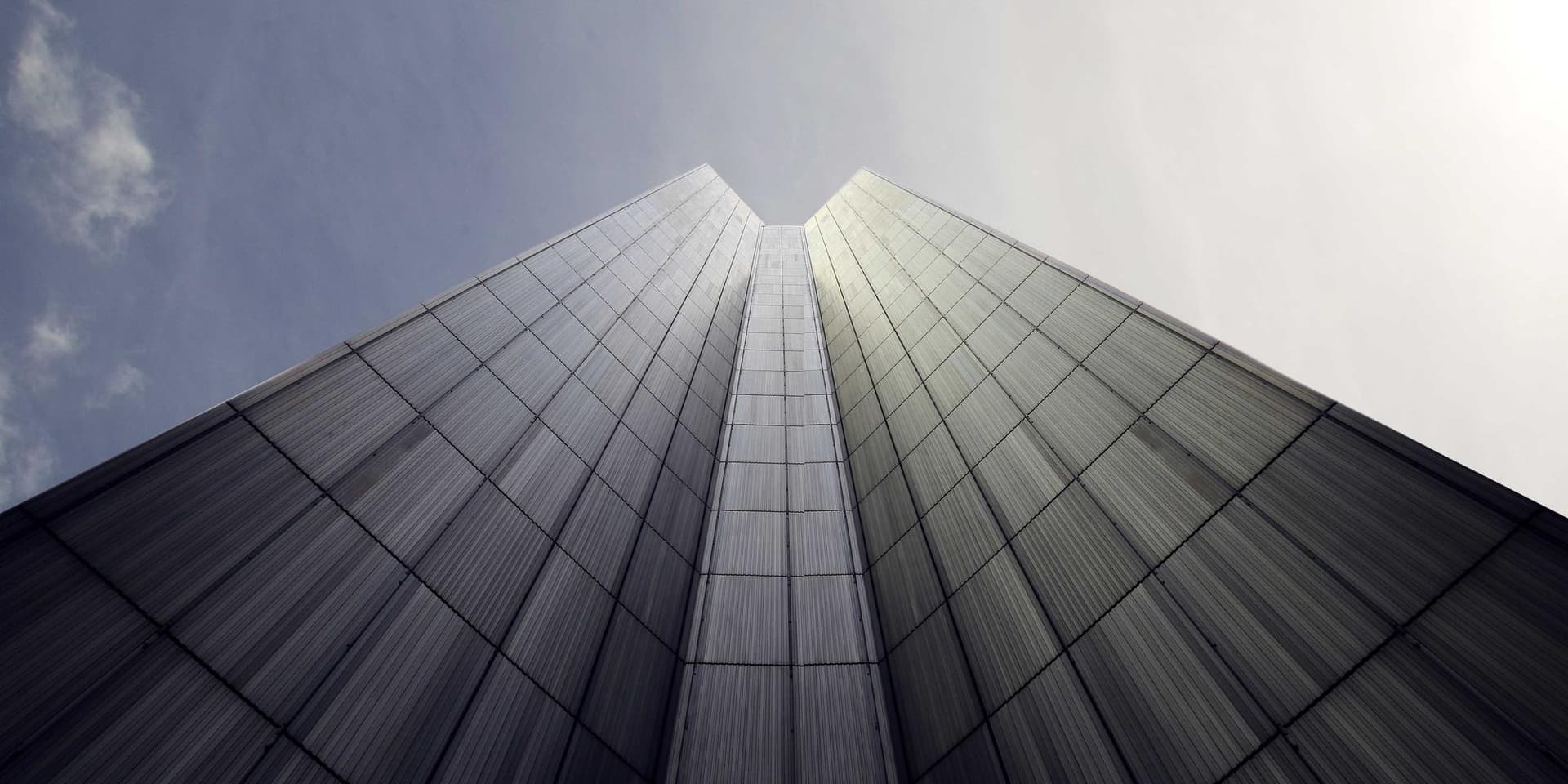 Fachada de vidro de um edifício vista de baixo, convergindo para a aresta central contra o céu