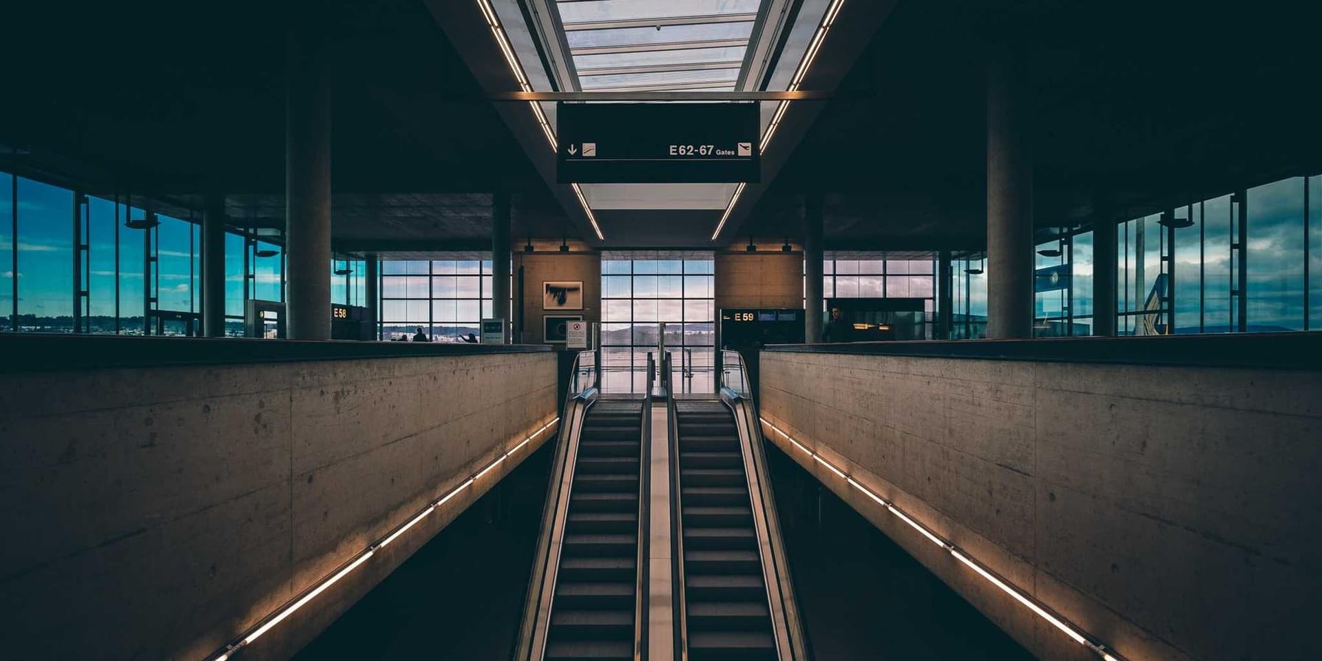 Escadas rolantes descendo entre paredes de concreto numa estação, com o saguão envidraçado ao fundo