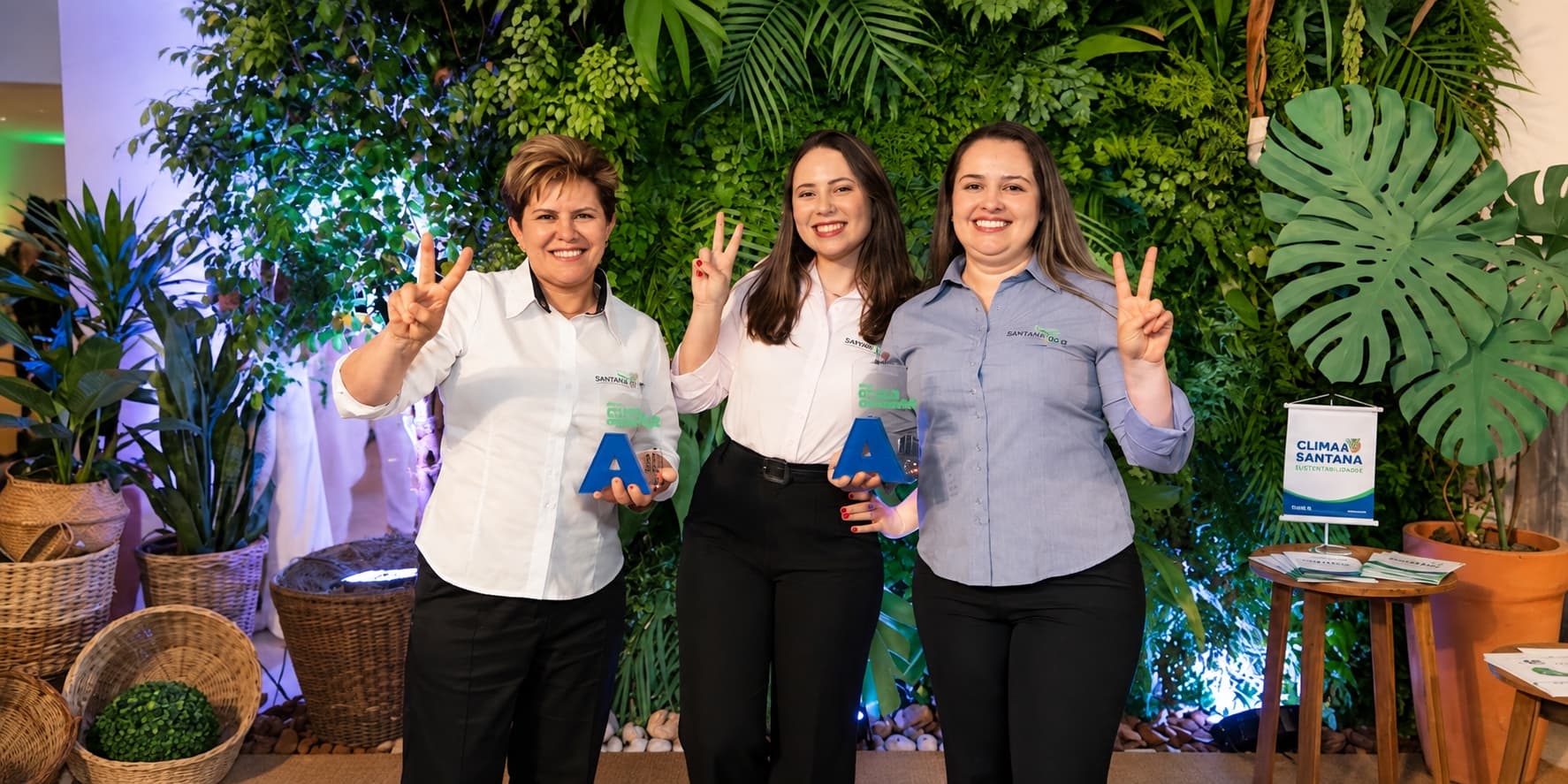 Três profissionais da Sustentabilis seguram os troféus do Clima Paraná Sustentável diante de um painel de plantas