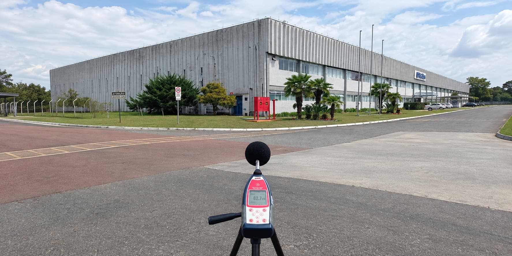 Medidor de nível de pressão sonora sobre tripé marcando 62,7 dB na via em frente a um galpão industrial